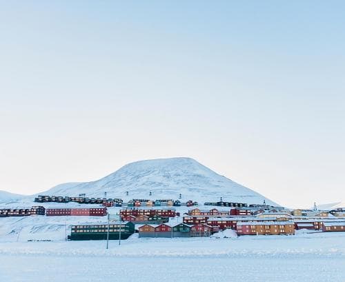 A serene Arctic settlement features colorful houses nestled against a snow-covered hill under a clear blue sky, ideal for travelers seeking unique polar landscapes and cultural experiences.
