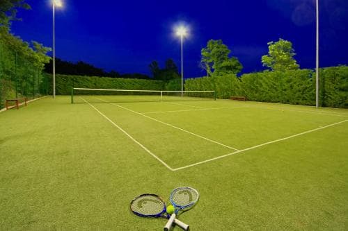 A serene night-time tennis court illuminated by floodlights features two rackets and a ball on the grass, ideal for sports enthusiasts seeking an evening game in a tranquil outdoor setting.