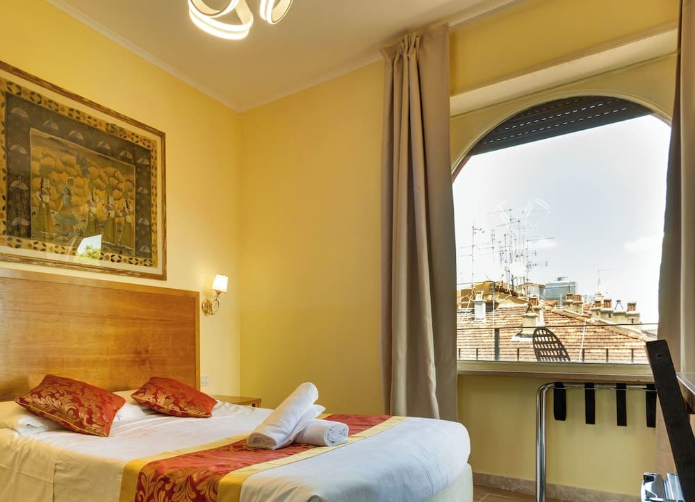 A cozy hotel room with yellow walls, a neatly made bed adorned with red accents, and an arched window offering a view of rooftops, ideal for travelers seeking comfort and charm in a cityscape setting.