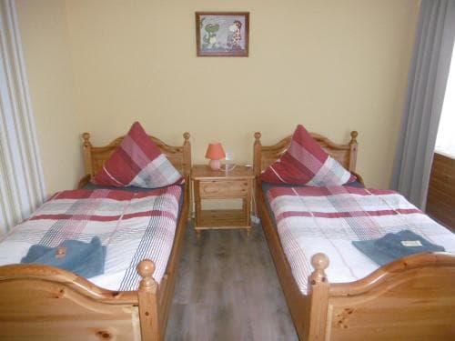 A cozy guesthouse room features two wooden single beds with plaid bedding, a small bedside table with a lamp, and a framed picture on the wall, ideal for travelers seeking simple and comfortable accommodation.