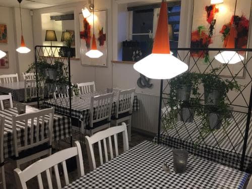A cozy indoor café features checkered tablecloths, white chairs, vibrant hanging lights, and artistic wall decor, creating an inviting atmosphere perfect for travelers seeking a charming dining spot.