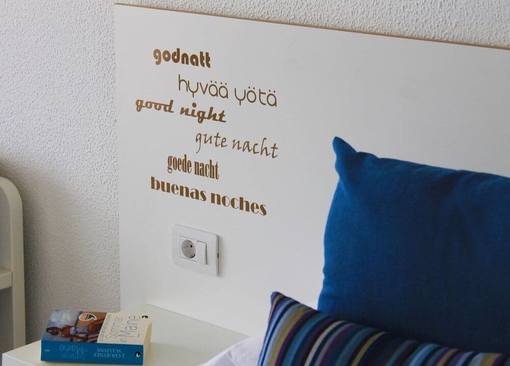 A cozy bedroom corner features a white headboard with "good night" written in multiple languages, complemented by blue pillows and a book on the bedside table, ideal for travelers seeking a comfortable and international ambiance.