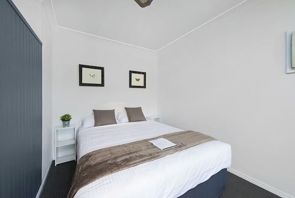 A cozy minimalist bedroom with white walls, neatly arranged bed with brown accents, framed wall art, and a small plant on the bedside table, ideal for travelers seeking a serene and tidy accommodation.