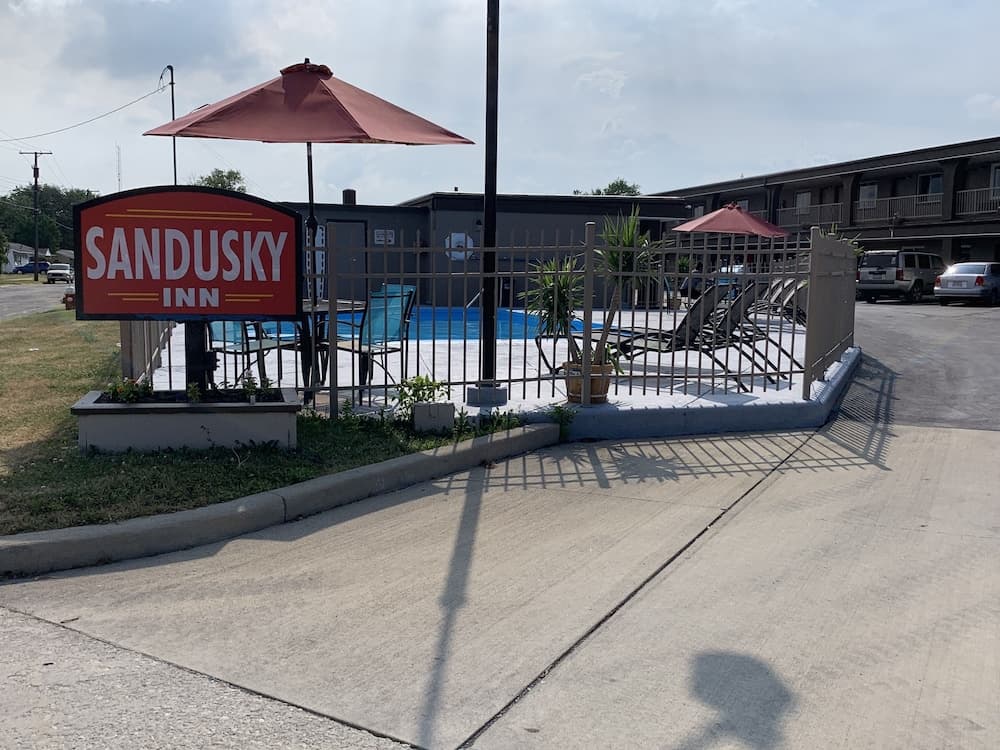 A welcoming motel entrance features the Sandusky Inn sign, a fenced pool area with lounge chairs under red umbrellas, and a calm suburban atmosphere ideal for travelers seeking relaxation.