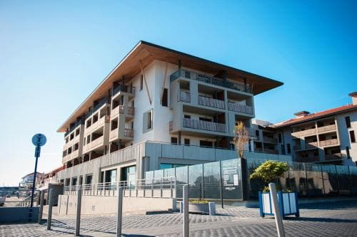 A modern multi-story apartment complex with wooden accents and balconies stands under a clear blue sky, surrounded by urban landscaping and street fixtures, ideal for travelers seeking contemporary accommodation in a vibrant city setting.