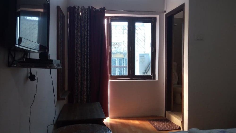 A cozy hotel room features warm wooden floors, a bright window with red curtains, a mounted TV, and an inviting ambiance perfect for travelers seeking comfort and relaxation.