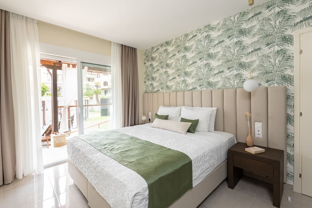 A serene hotel bedroom features a cozy bed with green accents against a tropical leaf-patterned wall, a balcony view of palm trees, and modern furnishings, ideal for travelers seeking relaxation and comfort.