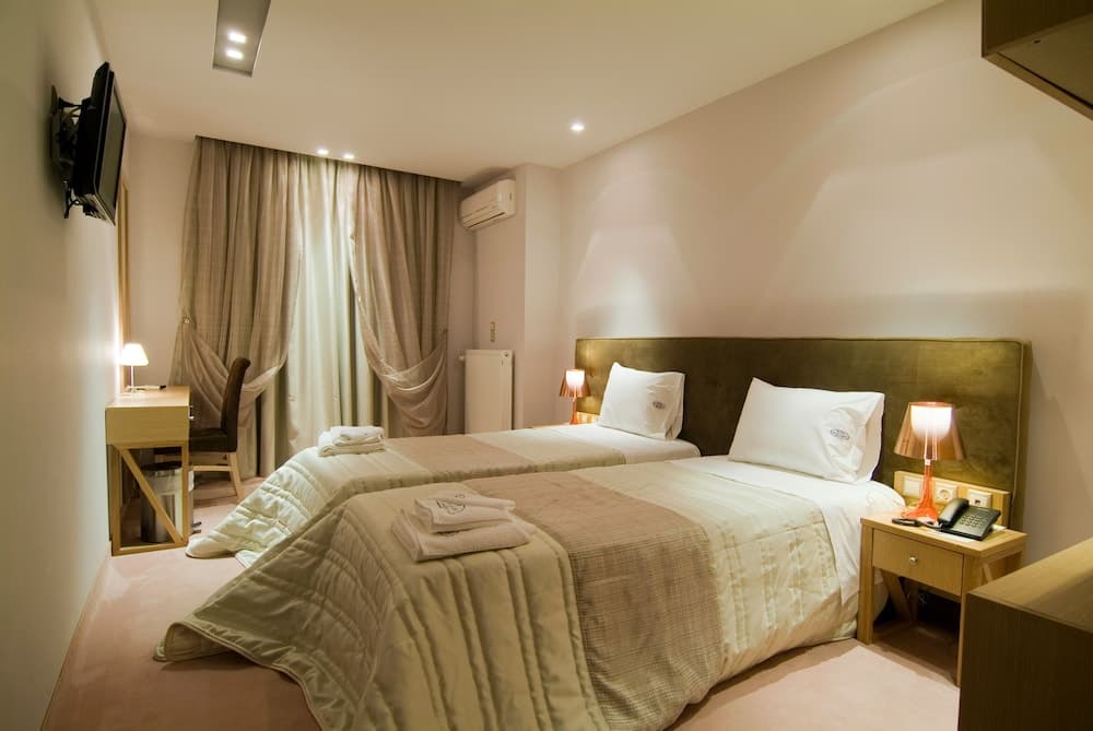 A cozy hotel room features twin beds with neatly folded towels, warm lighting from bedside lamps, a desk with a chair, and a wall-mounted TV, ideal for travelers seeking comfort and functionality.