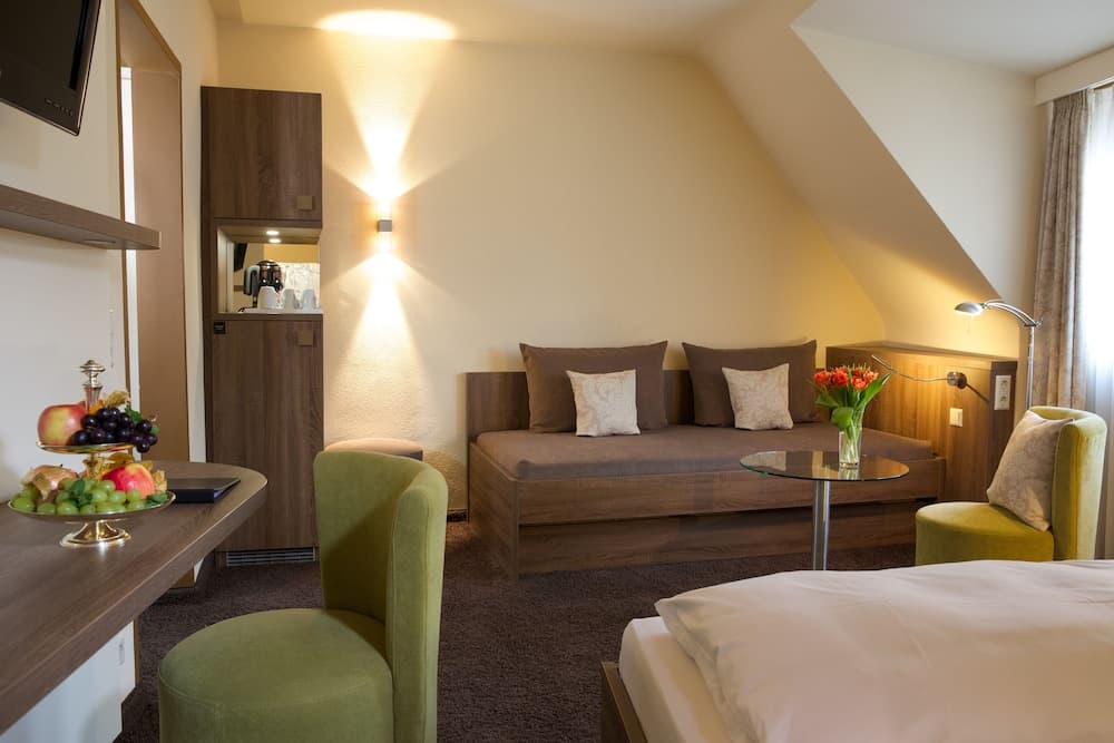 A cozy hotel suite features a warm-toned living area with a sofa, green armchairs, a fruit-filled centerpiece, and soft lighting, ideal for travelers seeking comfort and relaxation.