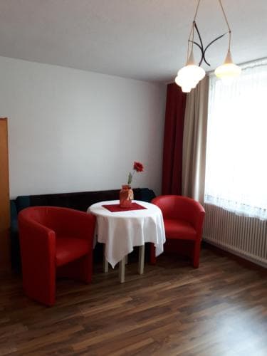 A cozy living room with red armchairs, a white tablecloth-covered table, and a modern chandelier creates an inviting atmosphere, ideal for travelers seeking a comfortable and stylish accommodation space.