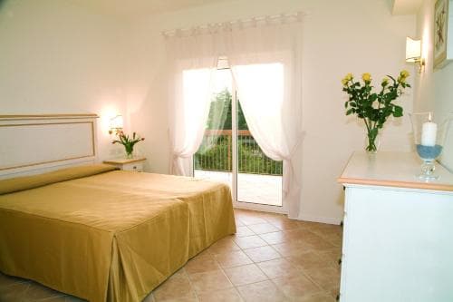 A cozy hotel room with a neatly made bed, tiled floors, and a balcony view of greenery, exuding a serene and inviting atmosphere ideal for travelers seeking relaxation.