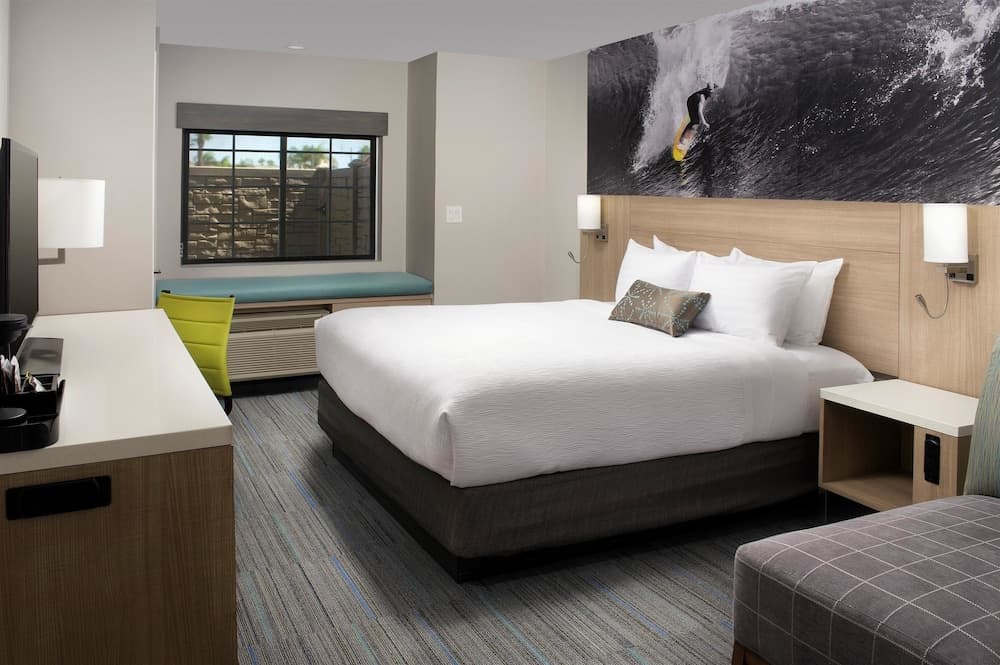 A cozy hotel room features a neatly made bed with a surfing mural above it, a vibrant yellow chair by the window, and a modern desk setup, ideal for travelers seeking comfort and functionality.