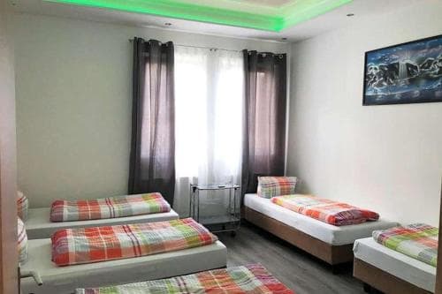 A cozy dormitory-style room with neatly arranged beds featuring colorful plaid blankets, soft lighting from green ceiling strips, and a framed picture on the wall, ideal for budget travelers seeking comfortable group accommodations.