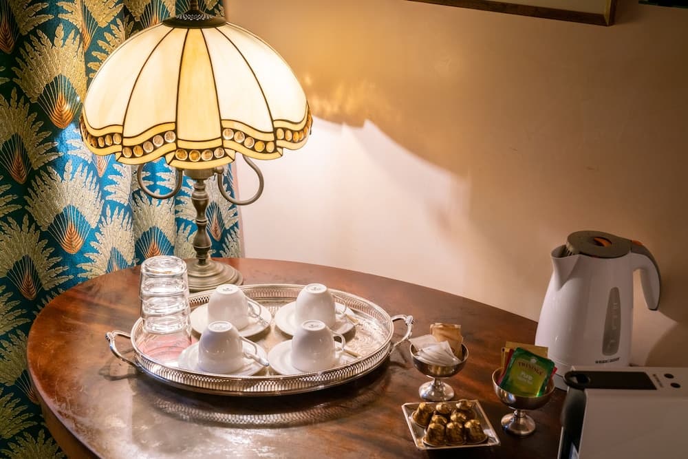 A cozy hotel room corner features a warmly lit table with a Tiffany-style lamp, elegant tea setup, snacks, and an electric kettle, ideal for travelers seeking comfort and charm.