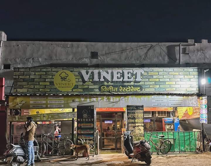 A bustling night scene at Vineet Restaurant in Sadar Bazaar, Meerut, featuring illuminated signage, parked scooters, bicycles, and a dog, evoking a lively local dining atmosphere ideal for travelers seeking authentic Indian cuisine.