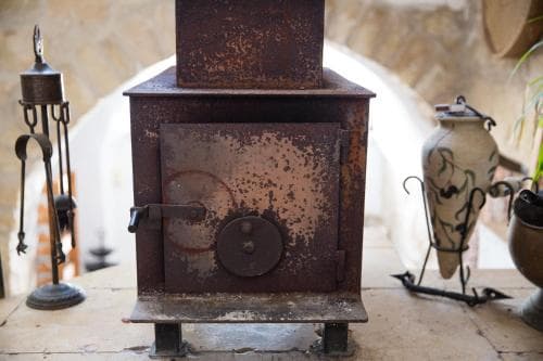 A rustic indoor setting features a weathered wood-burning stove surrounded by ornate metalwork and pottery, evoking a cozy, traditional atmosphere ideal for cultural heritage tourism.
