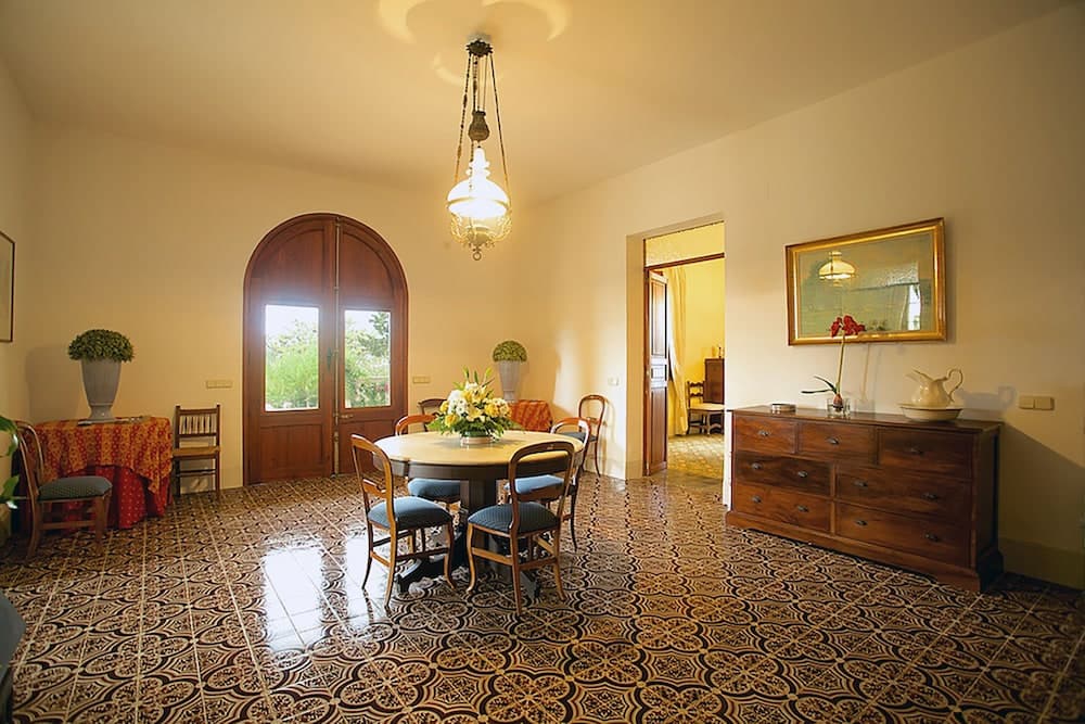 A cozy, warmly lit dining room with patterned tile floors, wooden furniture, and an arched doorway leading to greenery outside, ideal for travelers seeking a charming and inviting accommodation setting.