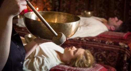A serene spa setting features individuals relaxing on patterned beds while a therapist uses a singing bowl, ideal for promoting wellness and stress relief during a tranquil retreat experience.
