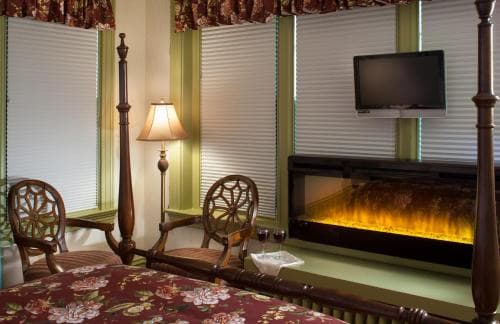 A cozy hotel room features a four-poster bed adorned with floral bedding, a warm fireplace, wooden chairs by the window, and a mounted TV, ideal for travelers seeking comfort and relaxation.