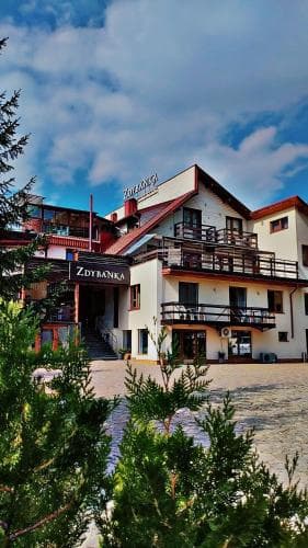 A charming multi-story hotel with balconies and a welcoming entrance is nestled amidst greenery under a partly cloudy sky, ideal for travelers seeking a cozy retreat in a serene mountainous area.