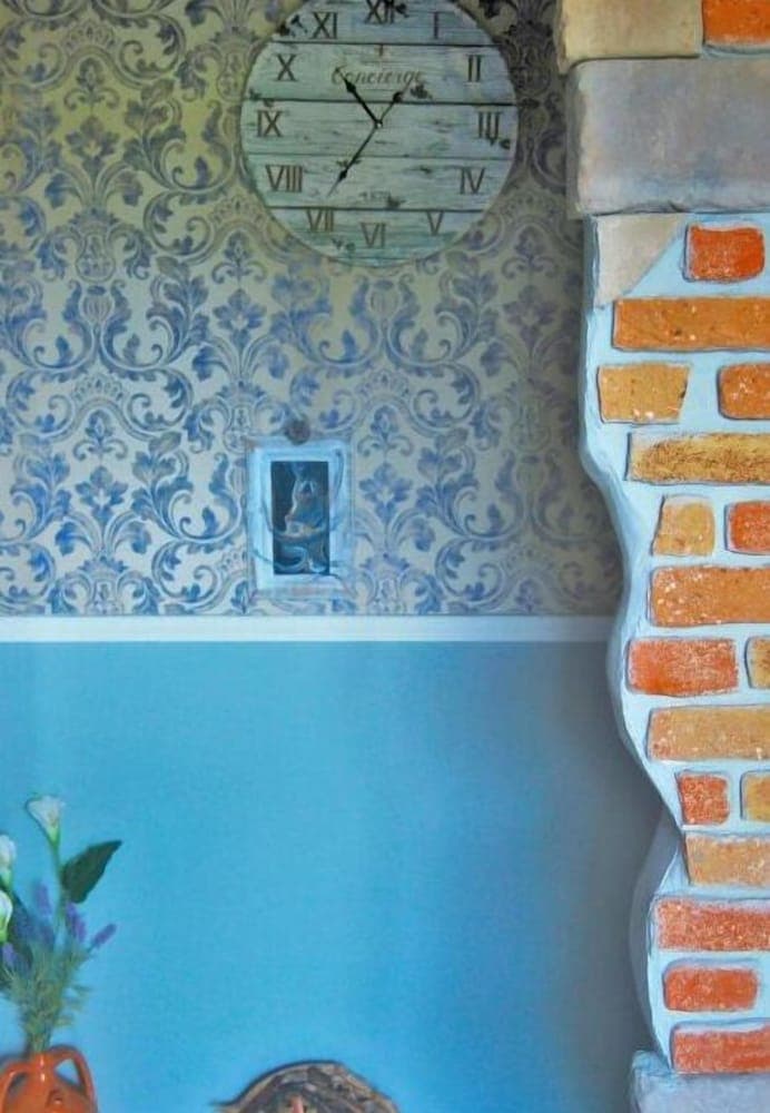 A charming indoor corner features ornate blue damask wallpaper, a rustic wooden clock, a brick-patterned staircase, and vibrant flowers in an orange vase, ideal for travelers seeking cozy, vintage-inspired accommodations.
