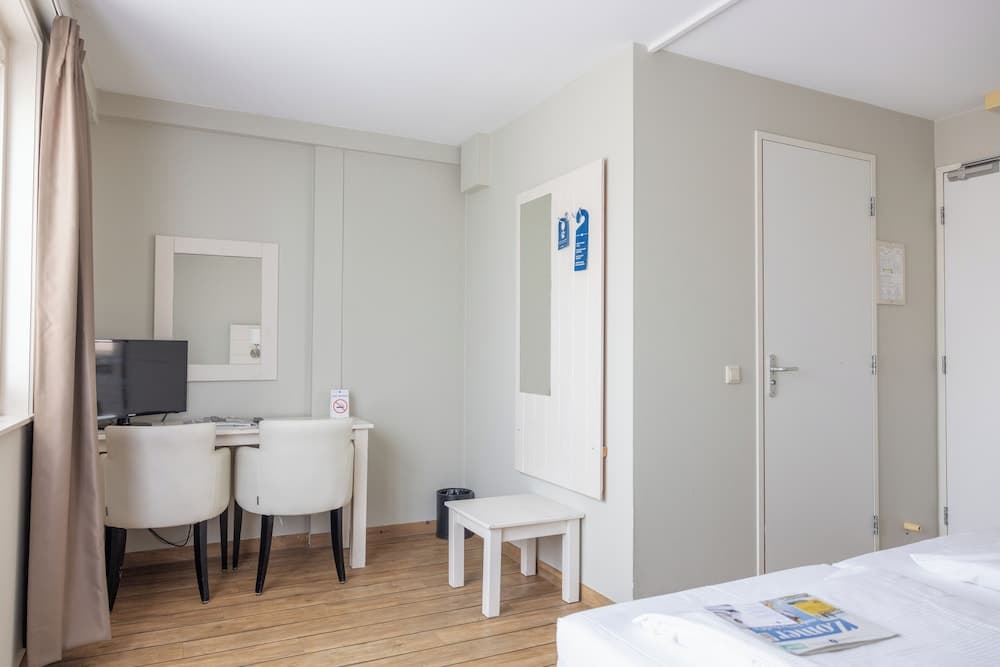 A cozy hotel room features a wooden floor, white furniture including a desk with chairs and a small bench, a mirror, and a bed with a magazine, ideal for travelers seeking comfort and simplicity.