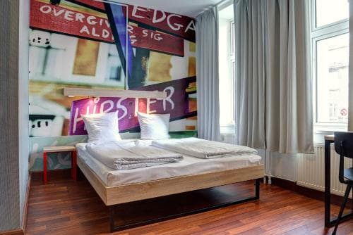 A cozy hotel room features a modern bed with vibrant graffiti-style wall art, wooden flooring, and natural light from large windows, ideal for travelers seeking a trendy urban retreat.