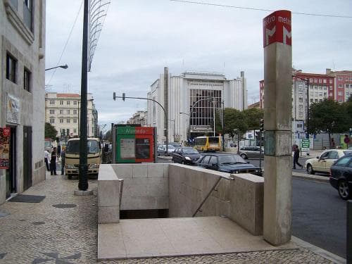 A bustling urban square features a modern metro entrance with a red sign, surrounded by classic European architecture, vehicles, and pedestrians, ideal for city exploration and public transportation access.