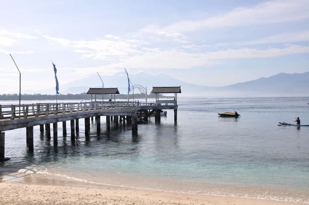 A serene beach scene features a long wooden pier with traditional pavilions extending into calm turquoise waters, framed by distant mountains under a soft blue sky, ideal for tranquil coastal getaways and photography.