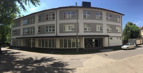 A modern multi-story building with gray and white panels stands under a clear blue sky, surrounded by greenery and a quiet street, ideal for travelers seeking accommodation in a serene urban setting.