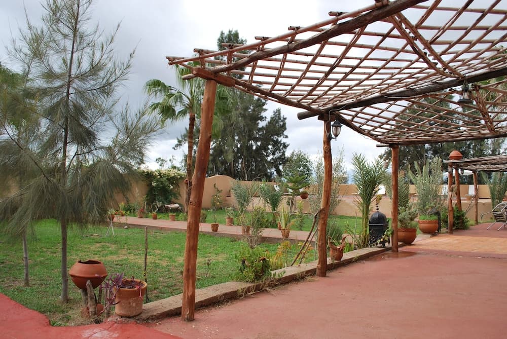 A serene outdoor patio with a rustic wooden pergola, terracotta pots, lush greenery, and a tranquil atmosphere invites travelers seeking relaxation in a natural setting.
