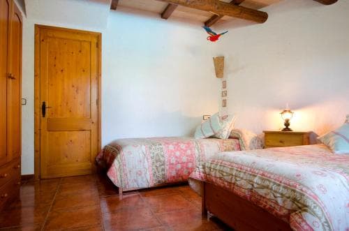 A cozy rustic bedroom with wooden beams, twin beds adorned in floral bedding, warm terracotta flooring, and a vibrant parrot decoration, ideal for travelers seeking a charming countryside retreat.