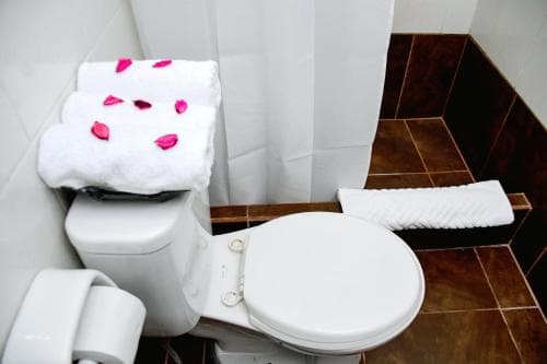 A cozy hotel bathroom features a toilet adorned with neatly folded towels decorated with pink rose petals, set against warm brown tiles and a white shower curtain, ideal for travelers seeking comfort and attention to detail.