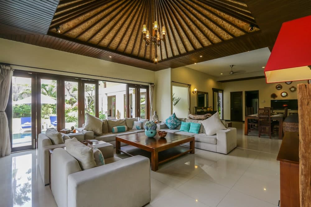 A luxurious villa living room with a high thatched ceiling, plush white sofas adorned with turquoise pillows, a wooden coffee table, and large glass doors opening to a serene outdoor pool area, ideal for travelers seeking a tranquil tropical retreat.