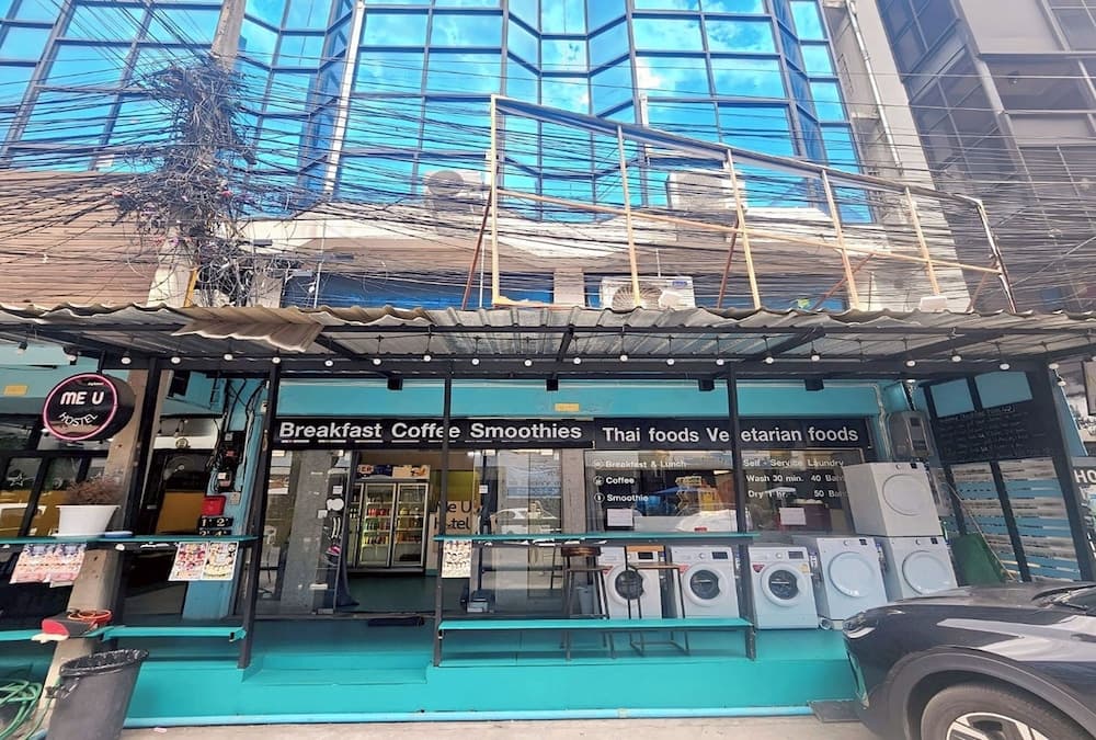 A vibrant street-side café with turquoise walls offers breakfast, coffee, smoothies, Thai foods, and vegetarian options under a bustling urban skyline, ideal for travelers seeking local cuisine and convenient amenities.