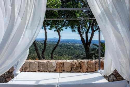A serene outdoor retreat features white draped curtains framing a stone wall and lush greenery, offering a tranquil view of distant hills, ideal for relaxation and luxury travel experiences.