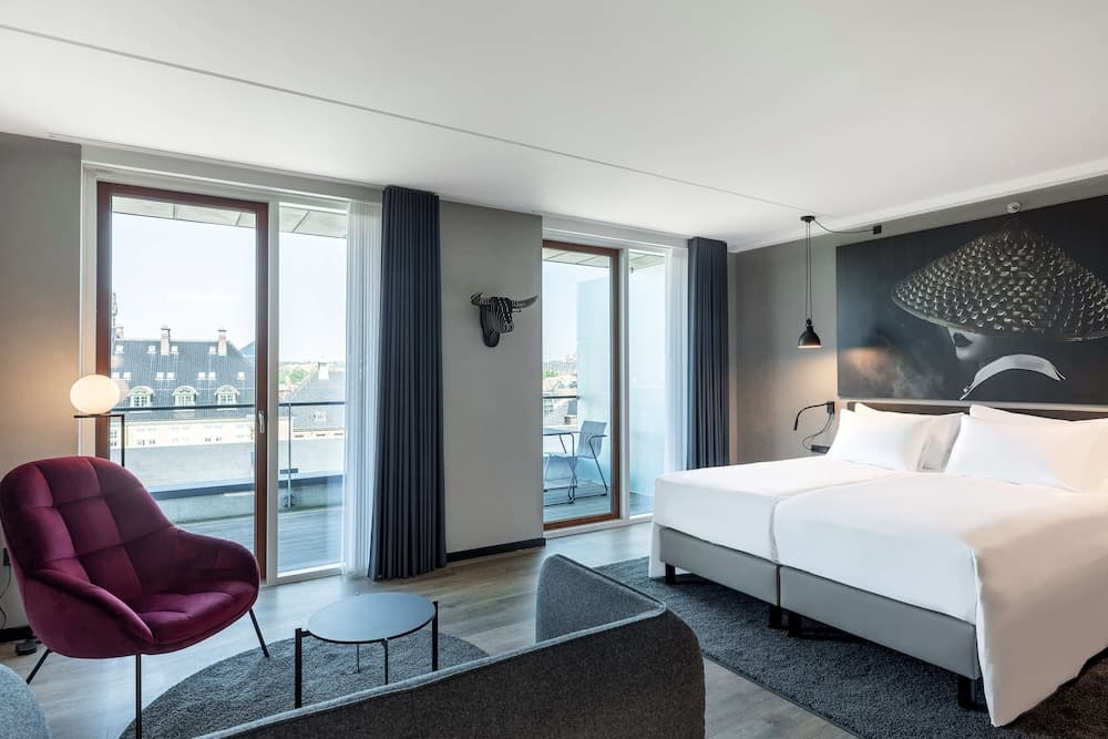 A modern hotel room with a cozy ambiance features a plush red armchair, sleek furnishings, and large windows offering a scenic view of rooftops, ideal for travelers seeking comfort and style.