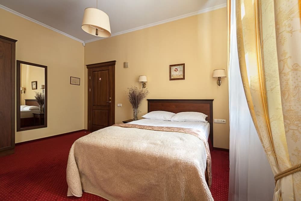 A cozy hotel room with warm yellow walls, a neatly made bed adorned with a beige blanket, elegant curtains, and classic furnishings creates an inviting atmosphere ideal for travelers seeking comfort and relaxation.