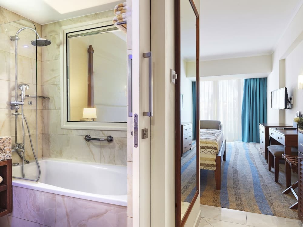 A luxurious hotel suite features a spacious bathroom with a marble bathtub and shower, transitioning into a cozy bedroom with a neatly made bed, vibrant blue curtains, and modern furnishings, ideal for travelers seeking comfort and elegance.
