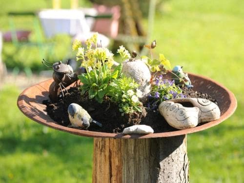 A charming miniature garden in a rustic metal bowl atop a wooden post, adorned with vibrant flowers, decorative stones, and playful figurines, creates a serene outdoor scene perfect for garden enthusiasts seeking inspiration.