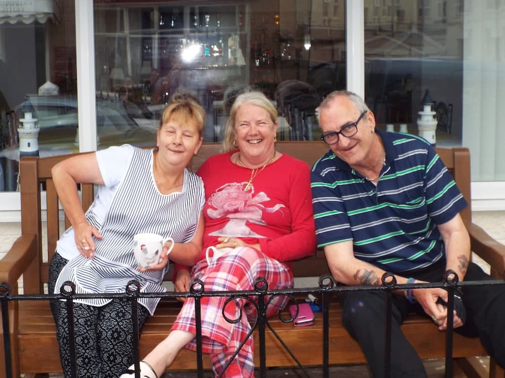 Three joyful individuals enjoy tea on a wooden bench outside a cozy café, with a cheerful atmosphere enhanced by their colorful outfits and the reflective window behind them, perfect for a relaxed getaway or family outing.