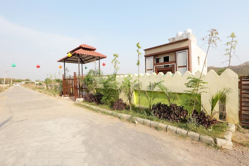 A serene roadside view of the Jhansi Castle Inn features a charming entrance gate adorned with colorful lanterns, lush greenery, and a modern building in the background, ideal for travelers seeking a peaceful stay in Jhansi, Uttar Pradesh.