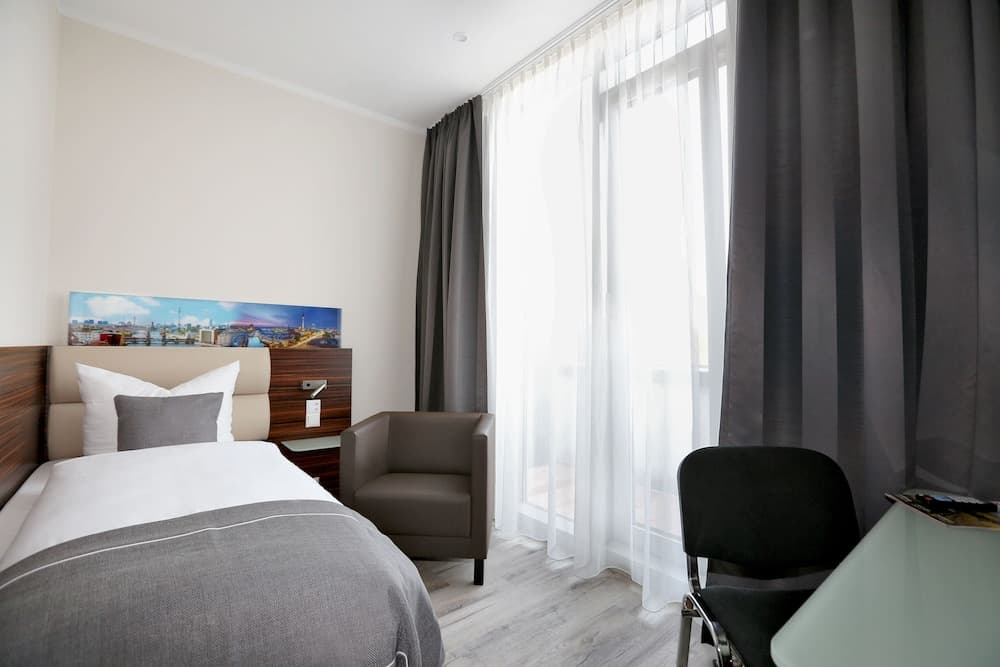A cozy hotel room features a neatly made bed, modern furnishings, and large windows with sheer curtains, creating a serene atmosphere ideal for business travelers seeking comfort and relaxation.