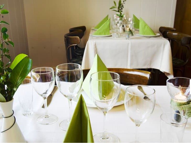 A cozy restaurant setting features neatly arranged tables with white linens, green napkins, and elegant glassware, creating an inviting atmosphere ideal for a romantic dinner or special occasion.