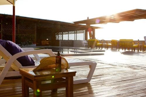 A serene poolside deck bathed in golden sunlight features lounge chairs, a wooden table with a bottle, and distant seating under a pergola, ideal for relaxing retreats or vacation photography.