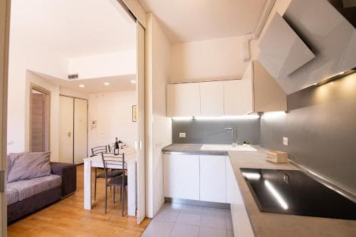 A cozy modern apartment kitchen with sleek white cabinets, stainless steel appliances, and an open layout leading to a dining area, ideal for travelers seeking comfort and convenience in urban accommodations.