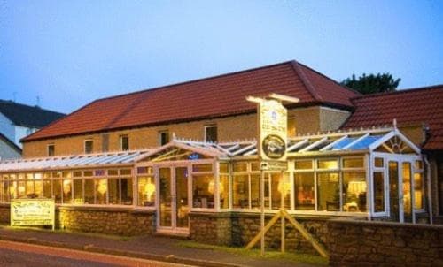 A cozy evening scene at a charming countryside pub with a glass conservatory extension, warmly lit interiors, and a welcoming signpost, ideal for travelers seeking a quaint dining or accommodation experience.
