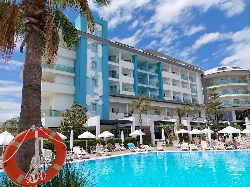 A vibrant resort scene featuring a turquoise pool lined with lounge chairs under white umbrellas, palm trees, and a striking blue and white hotel building, ideal for a relaxing vacation in Antalya, Turkey.