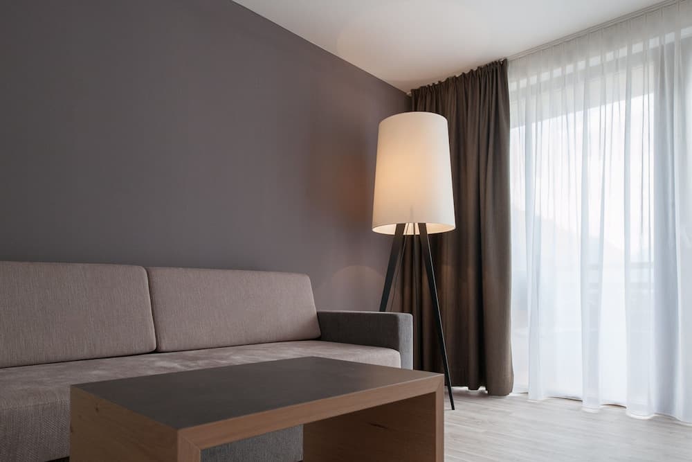 A cozy hotel room features a modern sofa beside a sleek coffee table, illuminated by a tall floor lamp near sheer curtains, ideal for travelers seeking comfort and style.