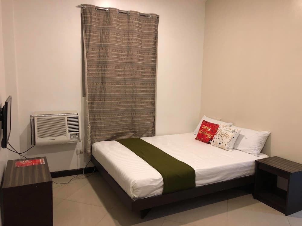 A cozy hotel room features a neatly made bed with festive pillows, a green throw blanket, wooden bedside tables, and a window with patterned curtains, ideal for travelers seeking comfort and simplicity.
